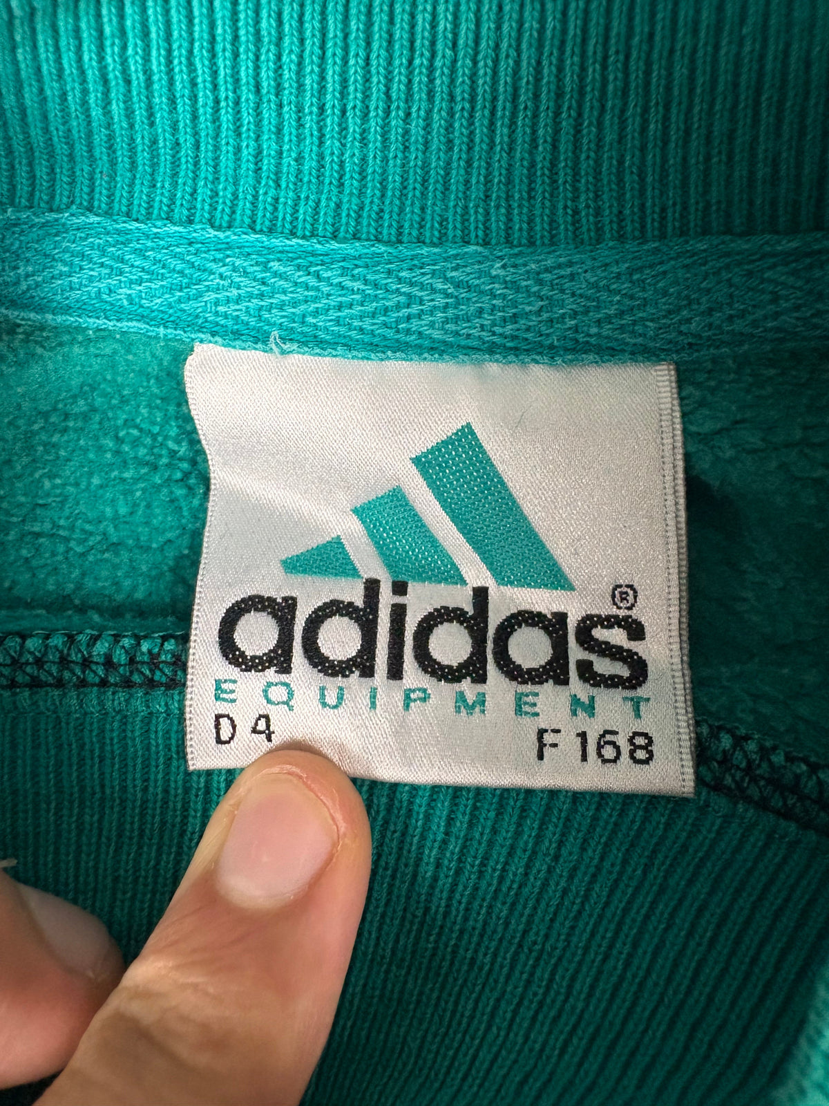 Innenetikett im Kragen des Adidas Equipment Sweaters mit türkis-schwarzem Equipment-Logo